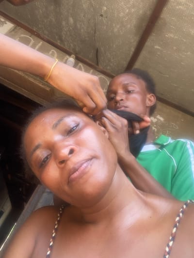 An image taken in Idiroko, Nigeria, depicts three individuals engaged in hair braiding under a corrugated roof. In the foreground, a dark-skinned female faces the camera, taking a selfie. She wears a multi-colored braided strap top. Behind her, a second dark-skinned female, wearing a green shirt with white arm stripes, focuses on braiding the first woman's hair, holding a section of dark synthetic hair. A third person's arm, adorned with a thin gold-colored bracelet, is visible in the upper left, also appearing to manipulate the hair near the first woman's scalp. The background shows a corrugated ceiling, likely made of asbestos cement sheets, supported by wooden beams. A small white object, possibly a light fixture or spray bottle, is affixed to a beam in the upper background.