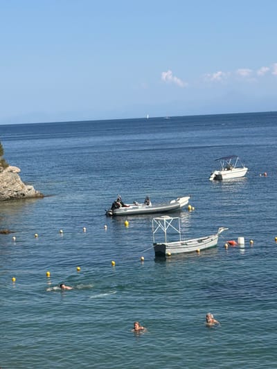 In the sea off the coast of Skiathos, Greece, on a clear day, several boats are anchored. A small, gray inflatable boat with an outboard motor is moored to the right. Further out, a white motorboat with a blue canopy is visible, also at anchor. In the foreground, a person is swimming in the water, while another person is partially submerged nearby. A line of yellow buoys marks a swimming area. In the background, a rocky outcrop with some vegetation juts into the sea, and the faint outline of mountains can be seen on the horizon. The sky is a clear, bright blue.