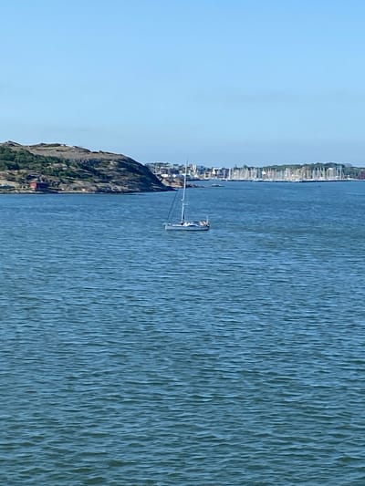 The image shows a body of water with a sailboat in the middle ground. In the background, a city skyline with numerous sailboats docked can be seen. A rocky, tree-covered hill is visible on the left side of the image, with a few red buildings at its base. The sky is clear and blue. The scene is consistent with a view of the archipelago or a harbor area in Gothenburg, Sweden, likely taken from a boat or ferry.