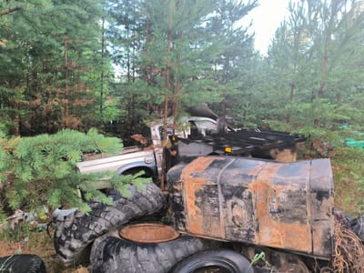 In a wooded area near a location identified as Гремучий, Russia, a scene of disarray is depicted. The foreground is dominated by several large, worn tires, some stacked and others lying haphazardly. Among these tires, a large, rectangular metal tank with significant rust and black paint is prominent. A circular metal object, possibly a lid or cover, rests on top of one of the tires.

Behind the tires and tank, parts of abandoned vehicles are visible. A silver pickup truck, appearing to be a Ford, is partially obscured by pine branches. Further back, another vehicle, possibly a van or truck, is visible with its roof and a portion of its white body showing. The environment is dense with pine trees, many of which have dried or brown needles, suggesting a natural setting that has become a dumping ground for discarded vehicles and materials. The overall impression is one of neglect and decay.