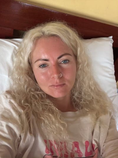 A woman with blonde, curly hair is lying on a white pillow in bed. She is looking directly at the camera. Her skin appears to be flushed, particularly on her cheeks and forehead. She is wearing a light-colored t-shirt with pink lettering that is partially visible. The headboard of the bed is dark red, and the wall behind it is yellow. The image was taken in Luang Prabang, Laos.