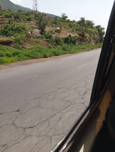 This media file captures a roadside view from inside a vehicle, likely taken in Rimin Ziem, Nigeria. The scene depicts a paved road with visible cracks, suggesting it's well-traveled. To the left of the road, a lush green hillside rises, dotted with various types of vegetation, including bushes and trees. Large rocks and boulders are interspersed on the hillside, adding texture to the landscape. In the background, a tall telecommunications tower is visible on the crest of the hill, indicating a developed area or a strategic location. The sky is clear and bright, suggesting a daytime scene with fair weather. The perspective from inside the vehicle, with a portion of its interior frame visible on the right, implies the image was taken while in motion, perhaps on a journey. The overall mood is serene and observational, focusing on the natural landscape and the infrastructure present in the region.