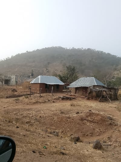The image presents an arid landscape in Jibam, Nigeria, under a hazy, pale sky. The terrain consists predominantly of dry, reddish-brown soil with sparse, desiccated vegetation and scattered rocks. A large, tree-covered hill, obscured by atmospheric haze, rises in the background.
In the mid-ground, two traditional single-story structures with reddish-brown earthen walls and corrugated metal roofs are situated. The structure on the left features two visible rectangular openings. The structure on the right has a dilapidated, open-sided wooden annex with a partial brush roof attached to its immediate right side. A small, rudimentary wooden fence stands in front of the left structure.
Further to the left background, a collection of more contemporary multi-story buildings, likely constructed of concrete, are visible; one features a black water storage tank on its roof. The immediate foreground shows a circular depression in the earth, edged with rocks, along with more scattered dry vegetation and debris. A partial view of a vehicle's side mirror is visible in the lower left corner. No persons or animate subjects are depicted in the scene.
