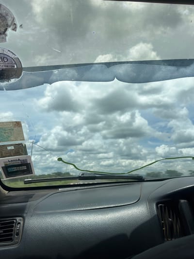 The image depicts an interior view from a vehicle, looking forward through its windshield towards an exterior landscape in Maji Moto/Naroosura ward, Kenya.

The windshield is visibly dirty, covered with dust and smudges, and exhibits several prominent cracks, notably a long, winding crack across the lower central area and another extending from the upper left. A dark, wavy sunshade strip is affixed to the upper interior edge of the windshield. Multiple official-looking rectangular stickers or permits are affixed to the upper left interior section, with one clearly displaying "Lami" and a QR code. A slender, bright green object, approximately 30-40 cm in length and consistent in appearance with a snake or vine, is positioned horizontally across the lower central part of the windshield, resting just above the dashboard. A windshield wiper blade is visible at the base of the windshield on the right side from the viewer's perspective.

The vehicle's dashboard, dark gray and textured, occupies the lower portion of the frame. The phrase "SRS AIRBAG" is embossed on the passenger side. Air vents are visible on both the left and right sides of the dashboard, which shows signs of dust accumulation.

Beyond the windshield, the exterior environment features a partly cloudy sky with large, white cumulus clouds set against patches of blue. The distant horizon reveals a flat landscape with sparse green vegetation, indicative of trees or bushes, consistent with a rural or savanna environment in Kenya. No individuals or specific actions are discernible.