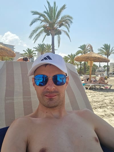 In S'Arenal, Spain, a man wearing a white Adidas baseball cap and mirrored sunglasses is reclined on a striped beach chair, facing the camera. The man is shirtless, and his skin is tanned. His sunglasses reflect the beach scene behind him, including palm trees and a beach umbrella. In the background, other beachgoers are visible lounging on chairs and walking on the sand. Buildings and more palm trees line the background, indicating a resort area. The sky is clear and blue, and the overall scene depicts a sunny day at the beach.