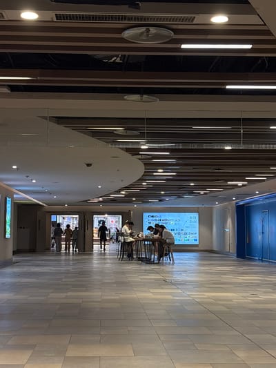 The image shows a modern, well-lit indoor public space, likely a shopping mall or commercial center in Heping District, China. The ceiling features a contemporary design with wooden slats and recessed lighting. In the background, a large digital screen displays what appears to be a directory or advertisement, with Chinese characters and various icons.

Several people are visible. In the middle ground, a group of four individuals are seated around a table, appearing to be engaged in conversation or using electronic devices. Their attire suggests casual wear. To the left, a group of three people are walking away from the viewer towards two open doorways, possibly shops or entrances. Another person is walking towards the right, away from the viewer.

The flooring is made of large, light-colored rectangular tiles, laid in a staggered pattern. The overall environment is clean and orderly, with an emphasis on modern architecture and lighting. To the far right, a section of a blue wall is visible, suggesting the presence of more commercial spaces or partitions. The scene is captured from a medium distance, providing a broad view of the area.
