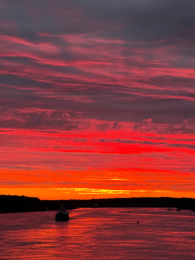 This image depicts a vibrant sunset over a body of water, with a silhouette of trees along the distant shoreline. The sky is filled with dramatic, striated clouds in hues of deep red, orange, and dark grey. The water reflects the intense colors of the sky, creating a shimmering, warm surface. In the foreground, a dark-colored vessel, possibly a freighter, is sailing away from the viewer on the water. Several smaller, indistinct objects, potentially buoys or other small boats, are scattered across the water. The overall scene suggests a serene and dramatic natural event taking place at dusk.