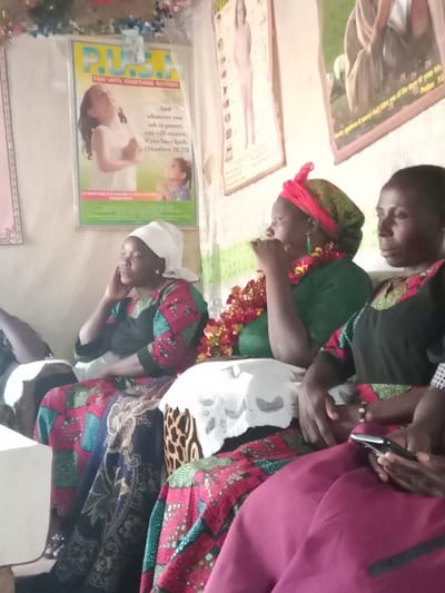 The image shows three women seated on a couch in a home in Kiminini, Kenya. The home is decorated with religious posters, including one with the phrase "PUSH: Pray Until Something Happens." The women are dressed in colorful clothing. The woman on the left is wearing a white headscarf and a red and black patterned dress. The woman in the middle is wearing a green dress with red trim and a red headscarf, and has a floral garland around her neck. The woman on the right is wearing a dark dress with a red and gold pattern and a white headscarf. The woman on the right is holding a black smartphone in her hand. The couch is covered in a patterned fabric and a white crocheted doily. The walls are decorated with posters and have a green and white pattern. The home is cluttered with items, indicating a lived-in space.