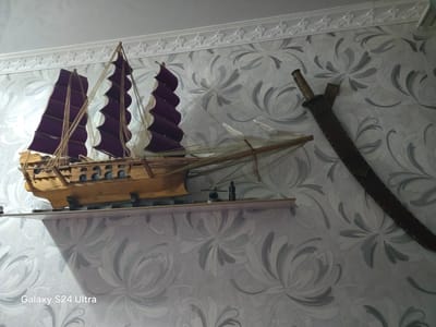 The image displays a wall with floral wallpaper and a decorative shelf. On the shelf rests a model of a wooden sailing ship with purple sails. To the right of the ship, a sword in a sheath is mounted on the wall. The scene is illuminated by a light source casting a shadow on the left side of the wall. The watermark "Galaxy S24 Ultra" is visible in the lower left corner. There are no people or actions depicted in the image. The location is specified as Novopavlovka, Kyrgyzstan.