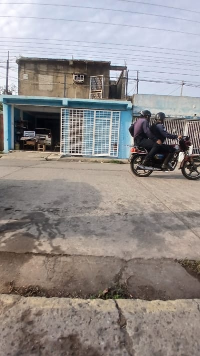 Two people are riding a motorcycle on a street in front of a building. Both individuals are wearing helmets. The building is painted blue and has a large garage door. Inside the garage, there are miscellaneous items, including what appears to be a vehicle, a desk, and boxes. The street is paved, and there is a curb on the right side. The sky is cloudy, and electrical wires are visible above the building. The location is Barcelona, Venezuela.