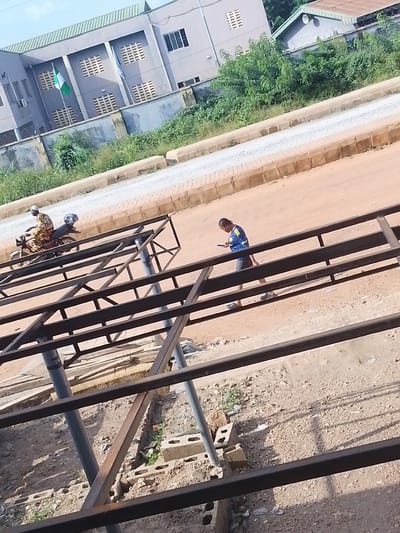 This media file captures an outdoor scene in Abeere, Nigeria. In the background, a light gray building with a green roof is visible. The building has multiple windows and ventilation grills, and two flags, one of which appears to be the Nigerian flag, are displayed. Adjacent to the building is a dense area of green foliage.
In the foreground, a person is walking on a dirt path, looking at their phone. They are wearing a blue shirt with yellow accents and dark shorts. The path is bordered by a gravelly area and some construction materials, including metal beams that form a skeletal structure, suggesting an ongoing construction project. Further in the background, a person is riding a motorcycle, wearing a patterned traditional outfit and a white cap. The motorcycle has a helmet on the back.
The scene appears to be set during the daytime, with the sun casting shadows on the ground. The weather seems to be clear. The overall impression is of a typical day in a developing area, with everyday activities like commuting and construction happening concurrently. No visible text is present in the image.