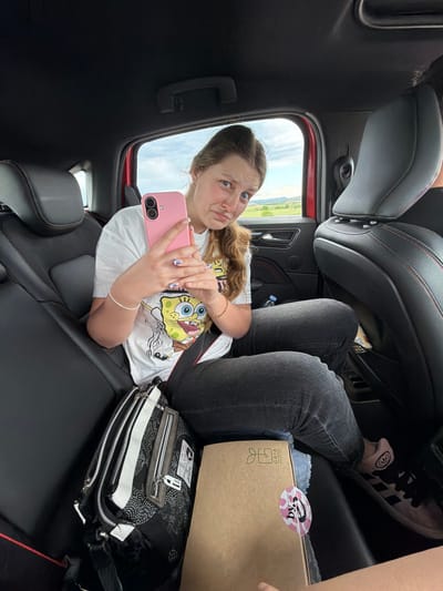 A young woman is inside the back of a red vehicle, possibly a car or SUV. She is lying sideways across the rear seat, facing the viewer. She has long blonde hair, fair skin, and blue eyes. She is wearing a white t-shirt with a cartoon character (Spongebob) printed on it, grey jeans, and sneakers. She is holding a pink smartphone in her hands, seemingly taking a selfie. The background visible through the car window shows a field or open space with a blue sky. There's a black and white patterned bag to her right. A Gaia packaging box is near her feet. The scene is within the interior of a car, but the specific location within Chokoba, Bulgaria, is not visually discernible.