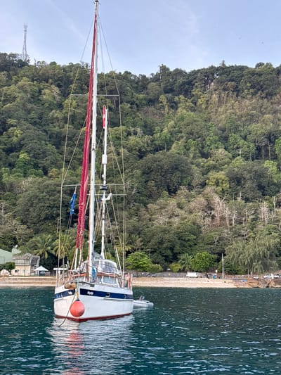 An anchored sailboat with a red buouy is docked in the turquoise waters of Christmas Island, Australia. The island's lush green hills are visible in the background. A communication tower is on the top of the hill. The sky is clear with a few birds flying. The sailboat has red and white sails furled and a blue and white hull. The name "Reykia" is visible on the stern.