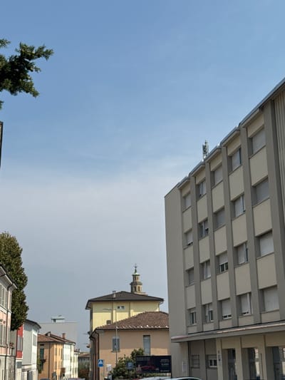 The image shows a street scene in Balerna, Switzerland, with a focus on buildings and the sky. In the foreground on the right is the side of a modern, multi-story building with a light beige facade and vertical gray accents. It has multiple windows with closed, slatted blinds. At the top of this building, a telecommunications antenna is visible. To the left of this building, a row of older, smaller buildings lines the street. These buildings have pitched roofs and varying facade colors, including pink, white, and yellow. A tall, mature tree with dark green foliage is on the left side of the street, partially obscuring the view of the buildings. In the background, a yellow building with a red-tiled roof is prominent, topped with a church steeple. Further in the distance, a more modern, white rectangular building is visible. The sky is a clear, pale blue with a few wispy clouds. There are no people or vehicles clearly discernible in the frame, and no overt actions or interactions are taking place.