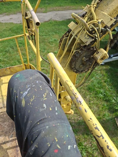 A ground-level, high-angle, outdoor shot shows a yellow, mechanical lift in an area with a grassy and dirt ground cover, ostensibly in the city of Zhabinka, Belarus. The lift’s structure is primarily tubular, with peeling yellow paint. The mechanical components, including a chain and various cables and hoses, are also painted yellow. A leg covered in black pants with paint splatters is positioned to the right, near the lift’s support. The background is a mix of green grass and a dirt pathway.