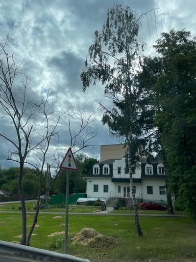 The image captures a scene in Semyonovskoye, Russia, under a cloudy sky. In the foreground, bare tree branches partially obscure a triangular road sign depicting a traffic light. Beyond the sign and branches, a white two-story building with dark gray trim is visible. A red car is parked beside the building. The scene includes green lawns and various trees, some of which partially block the view of the building. A section of a metal railing is visible in the lower right corner of the frame.