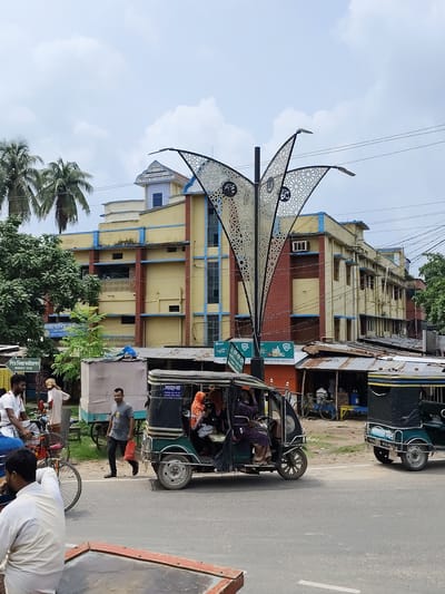 The image depicts a street scene in Benapole, Bangladesh. In the foreground, a man wearing a white shirt is partially visible, facing away from the camera. To his left, another man on a red bicycle is also facing away. Behind them, a green auto-rickshaw, a common mode of transport in Bangladesh, is seen with several passengers inside. The auto-rickshaw is parked on the side of a paved road, which appears to be a main thoroughfare.

Further down the road, another auto-rickshaw is parked on the right side. The background features a yellow building with blue trim, likely a government or institutional building. In front of this building are several shops with tin roofs, indicating a busy commercial area. Trees, including palm trees, are visible, suggesting a tropical or subtropical climate. A decorative lamppost with an intricate metal design stands prominently in the center of the scene, illuminating the area. The sky is partly cloudy. The overall atmosphere suggests a typical day in a bustling Bangladeshi town.