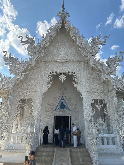 The image displays the highly ornate, all-white exterior of a temple in Ban Rong Khun, Thailand, under a clear blue sky with scattered white clouds. The structure features intricate stucco work, mirrored glass accents, and multi-tiered roofs adorned with numerous mythical creature sculptures, including prominent naga figures. A central, arching entryway is visible, with a dark rectangular door below a stylized blue and white triangular motif. Steps lead up to this main entrance.
Approximately eight individuals are present near the entrance. A group of five to six people, dressed in various casual attire including dark shirts, light tops, and a long dark dress, are gathered on the steps, with several appearing to enter the dark doorway. One individual in white pants and a white short-sleeved top with a shoulder bag stands to the immediate right of the entrance. In the lower left foreground, a person wearing a dark baseball cap and a patterned shirt is holding a smartphone vertically, aiming it towards the temple, suggesting they are capturing a photograph or video. The scene is brightly lit by natural sunlight, casting minimal shadows directly beneath some architectural elements.