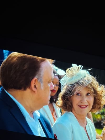 The image shows a medium shot of a woman wearing a light blue dress and a decorative hat with netting. She is smiling and looking to her right. To her left, partially visible, is a man with short brown hair and a suit jacket over a light blue shirt. In the background, out of focus, is another woman with darker skin and dark hair, wearing orange. The scene appears to be outdoors, possibly at an event, with greenery visible in the background. There is no indication of the location being in Ventspils, Latvia.