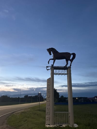The image depicts a twilight scene in Nikolskoye, Russia, featuring a monument of a horse on a pedestal. The monument is positioned near a road with streetlights lining its side. The sky is a deep blue with scattered clouds, indicative of dusk. The horse sculpture is dark in color and poised with one leg raised. The pedestal is a light gray structure with vertical text that appears to read "КОНЕЗАВОДСКИЙ" which translates to "Stud Farm". The road is paved and curves to the left, bordered by green grass on both sides. In the background, trees and residential buildings are visible. There are no people or animals in the immediate vicinity of the monument or on the road. The overall atmosphere is calm and serene, characteristic of the transition from day to night.