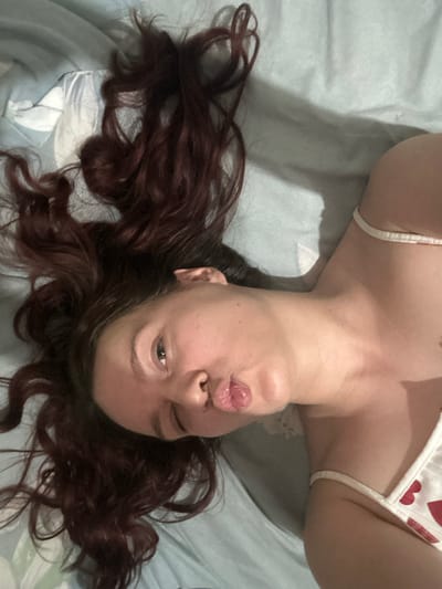 The media file shows a close-up, upside-down shot of a person lying on their back on a blue and white patterned bed. The person has long, dark, wavy hair that is spread out around their head. They are wearing a white top with a pattern of red hearts and a thin strap. Their face is turned towards the camera, with their lips pursed in a kissing gesture and one eye winking. The lighting appears to be artificial, suggesting the photo was taken indoors, likely in a bedroom setting. The overall mood conveyed by the image is playful and intimate. There is no visible text in the image. The location context of Bishkek, Kyrgyzstan, does not provide any specific visual cues within the image itself.