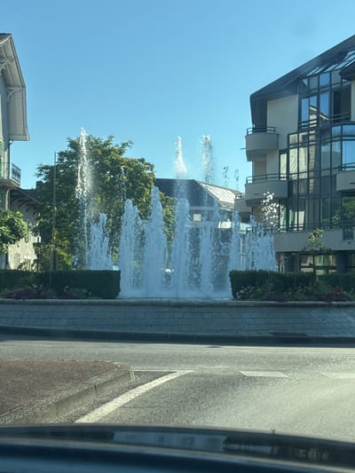 The image shows a fountain in the center of a roundabout in Divonne-les-Bains, France. The fountain has multiple streams of water spraying upwards, illuminated by the sun. Around the fountain is a circular area with manicured hedges and some flowering plants. In the background, there are buildings on either side of the roundabout. The building on the right is modern with large glass windows, while the building on the left appears older with a more traditional architectural style. The sky is clear and blue, indicating a sunny day. The foreground shows a section of the road, with white lane markings visible, suggesting this is a street view.