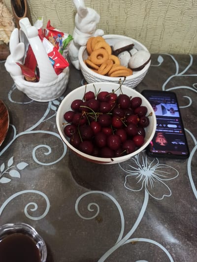 The image depicts a table setting with various snacks and a mobile phone. In a white ceramic basket shaped like a bunny, several individually wrapped candies are placed. Next to it, a white bowl contains ring-shaped cookies and meringue cookies with chocolate filling. In the foreground, a white bowl is filled with fresh cherries. To the right of the bowl of cherries, a smartphone is lying screen-up, displaying a social media feed with images of people. In the bottom left corner, a small glass dish with a dark liquid, possibly coffee or tea, is visible. The table surface has a dark brown pattern with light blue floral and swirling motifs. The background shows a textured wall. The scene is likely indoors. There is no visible indication of the city of Verkhnii Lars, Russia, or any specific location.