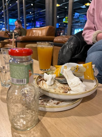 A cluttered table holds remnants of a meal. There are two dirty plates with leftover food and used tissues, along with a snack pack of what appears to be banana-flavored snacks. A glass of orange juice sits beside the plates. An empty glass bottle with a red lid is positioned in the foreground, its label partly visible. People are visible in the background, seated in lounge chairs. The scene is set in what looks like an indoor restaurant or lounge, with dark decor and blue lighting visible through the windows.