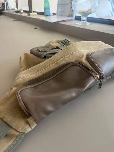 A tan canvas bag with brown leather accents is placed on a light-colored surface. The bag has multiple zippered compartments. In the background, a clear glass bowl filled with colorful small objects, possibly beads or marbles, sits on a windowsill. A green bottle of San Pellegrino water is also visible on the windowsill, along with a framed picture and a trophy. The environment appears to be indoors, likely in a modern building, given the clean lines of the furniture and the window. This scene is located in Germasogeia Municipality, Cyprus.