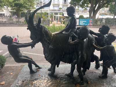 A bronze sculpture is depicted in a public outdoor setting in Vevey, Switzerland. The sculpture features a large, dark-patinated ibex or goat-like animal with prominent curved horns, engaged with five nude child figures. One child is seated on the animal's back, facing forward. Another child pulls the animal's head from the front left, leaning back with extended arms. Two children push the animal from its right side, while a fifth child on the left interacts with the animal's midsection. The sculpture rests on a textured bronze base, with scattered dry leaves on the ground around it.

In the immediate background, a paved surface, likely a sidewalk or street, is visible. Further back, to the left, is an outdoor dining area with white tables and chairs under awnings, fronting a multi-story building with balconies. A lamppost and a large tree with green foliage are present. Towards the right background, a blue and orange banner or poster is displayed on a pole, advertising "BUDDHA BAR BEACH" and "GRAND HOTEL DU LAC," identifying the specific vicinity of the Grand Hotel du Lac. A white sign with a red circle and diagonal line is visible to the left of the sculpture.
