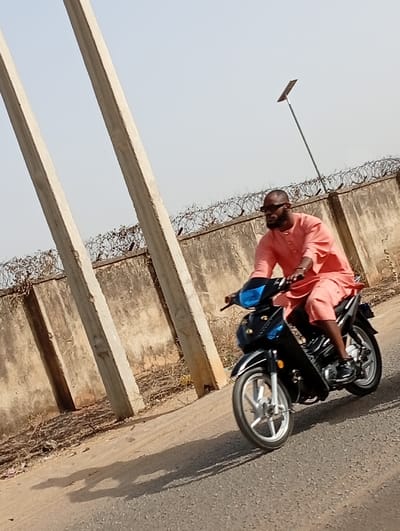 A male individual is observed riding a black step-through motorcycle on an asphalt road in Gayawa, Nigeria. The rider, who is bald with a short beard, wears dark sunglasses and a long-sleeved, knee-length, peach-colored traditional garment. The motorcycle features silver-colored five-spoke wheels. In the background, two light-colored concrete utility poles lean diagonally, originating from the left foreground. Behind these, a textured concrete wall runs horizontally across the midground, topped with multiple coils of barbed wire. Further back and to the right, a slender pole topped with a solar panel is visible above the wall. The sky is clear and light-colored, indicating daylight, and distinct shadows are cast onto the road surface, suggesting a high sun angle.
