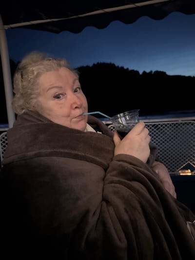 A woman is sitting outside, wrapped in a brown blanket, and drinking from a clear plastic cup with a blue straw. She has blonde curly hair and is looking towards the camera. The background is dark with a blue sky and silhouetted trees. The scene appears to be outdoors at dusk.