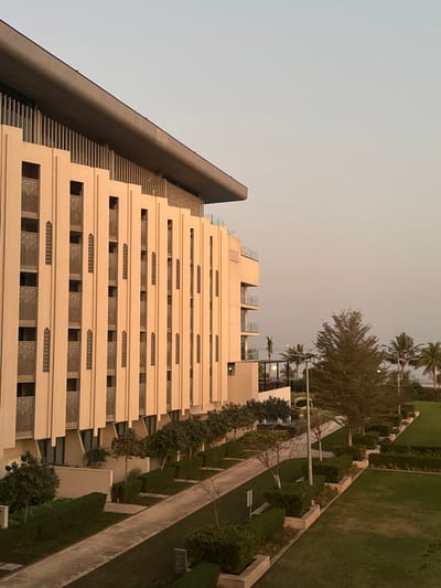 A multi-story building with a unique tiered design dominates the frame, its beige facade accented by rectangular openings. A dark gray structure adjoins the building, featuring stepped elements. Adjacent to the building is a meticulously landscaped garden with a pathway, lush green grass, and rows of shrubs and trees. The sky above is a pale gradient of light colors. The image is rotated.