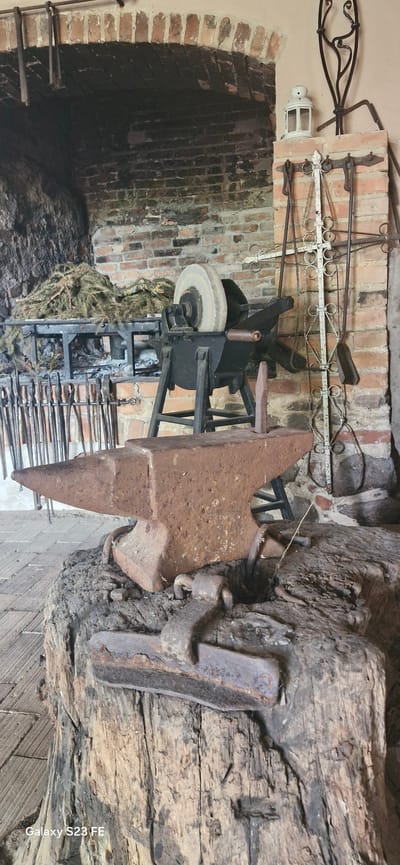 This is a medium shot of a rustic blacksmith's workshop in the city of Pakruojis, Lithuania. The scene is dominated by a large, weathered anvil resting on a thick, cracked wooden stump. Behind the anvil, a grinding wheel is mounted on a black metal stand, with a pile of evergreen branches behind it. To the right of the grinding wheel, various blacksmith tools hang on a decorative metal rack. The background features a large brick fireplace and a brick wall, suggesting an authentic historical setting. The lighting is dim, casting shadows and highlighting the textures of the metal and wood. The overall impression is one of a preserved historical craft or a staged display.