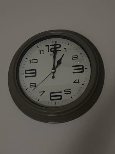 A wall clock is displayed against a neutral-colored wall. The clock has a round, dark frame and a white face with black Arabic numerals. The hour hand is pointing slightly past the 12, and the minute hand is pointing at the 3. There is a small logo that says "GAUDI" in the center of the clock face. The image appears to be taken in the city of Kocasinan, Turkey, but no specific location within Kocasinan is identifiable from the image. The scene is static and does not contain any people or discernible actions. The lighting is dim, suggesting it may be indoors or during low-light conditions.