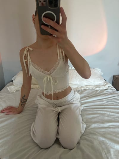 A person is kneeling on a bed, holding a phone to take a mirror selfie. The person is wearing a white camisole with lace trim and white pants. They have a snake tattoo on their left arm and a belly button piercing. The bed is covered with white sheets. The background is a wall with a soft, diffused light. The scene appears to be indoors, in a bedroom. There is no indication of the city of Ternopil, Ukraine in the image.