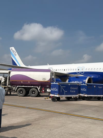 An Indigo Airlines airplane is parked on a tarmac. A fuel truck with a purple and white tank is positioned next to the plane. Several blue baggage carts with "Indigo Ground Force" written on them are also present. A person in a light blue shirt and dark pants stands to the left of the frame. The sky is mostly clear with some scattered clouds. The scene appears to be at an airport in Vikram Nagar, India.