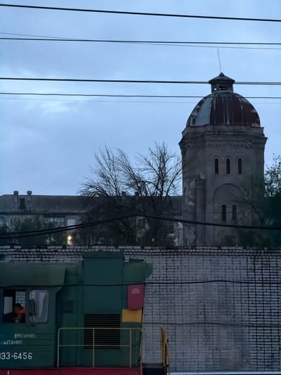 The image shows a water tower, a brick wall, and a green and red train locomotive in Rtishchevo, Russia. The tower has a dome-shaped top and several arched windows. Bare tree branches are visible in front of the tower and brick wall. Power lines run across the sky. The train locomotive has the number "933-6456" printed on its side. A person is visible through the train cab window. The overall lighting suggests it is dusk or dawn.
