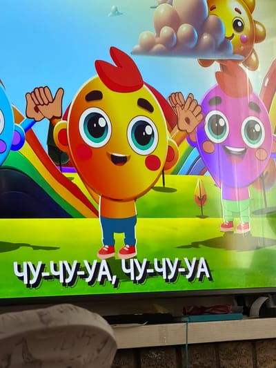 The image shows a television screen displaying a cartoon with several colorful characters. The characters appear to be variations of teddy bears or animal-like figures with large eyes. The foreground character is yellow and orange with a red "horn" on its head, wearing blue pants and red shoes. It is giving a thumbs-up. Other characters are purple and yellow, arranged in the background, with a rainbow behind the foreground character. Text is visible on the right side of the screen. The image is taken in an indoor environment, possibly a home, as indicated by the wall with brick-like wallpaper on the right side. The location in the city of Bearpark, United Kingdom cannot be confirmed, as the image does not contain any identifiable location markers or landmarks.
