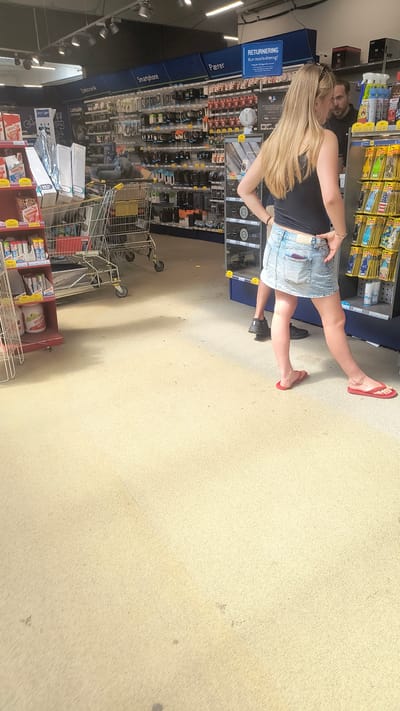 In a store in Køge, Denmark, a woman with long blonde hair stands with her hands on her hips, wearing a black tank top and a denim skirt. A man with a beard is visible behind her, looking towards the woman. They are standing in an aisle of a retail store, with shelves of electronics and other merchandise visible in the background. A shopping cart is also present in the aisle. The floor is a light-colored carpet.