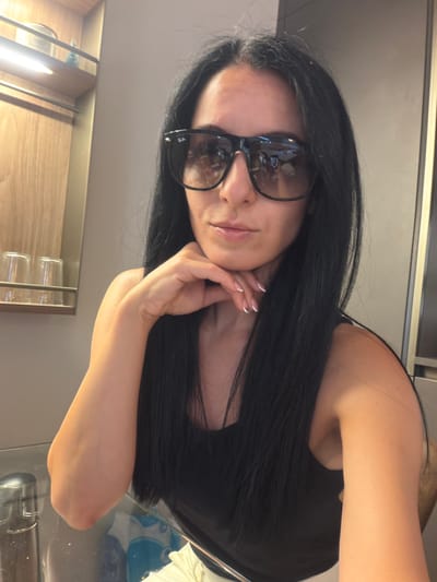 A medium shot captures a woman with long, dark hair and sunglasses, leaning her chin on her right hand. She wears a black tank top and white pants. The background features a wooden shelf with items on it, a grey wall, and a glass table. The image appears to be taken indoors, possibly in a hotel room or a similar setting, and no specific location within Buccinasco, Italy, is discernible from the visual information provided.