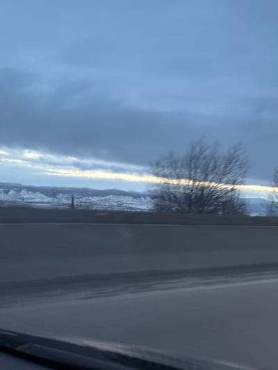 The photo shows a view through a window. Outside, a landscape unfolds under a cloudy, blue sky. The ground is covered in patches of snow, and bare trees stand silhouetted against the horizon. A thin band of bright light illuminates the landscape. A small figure of a person can be seen in the landscape. The window frame is visible on the right side of the image.