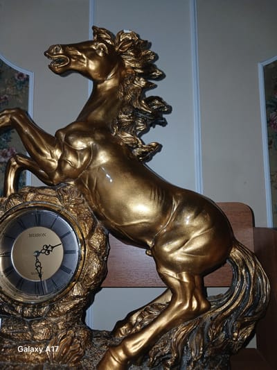 The image shows a close-up of a decorative gold-colored clock featuring a rearing horse sculpture. The horse is depicted in motion, with its mane and tail flowing. The clock face is round with Roman numerals and has "MIRRON" and "QUARTZ" visible on it. The clock is part of a larger decorative piece that is golden and appears to be made of resin or metal. The background consists of a wall with floral wallpaper at the bottom and a lighter-colored wall with molding above. A wooden shelf is visible on the right side. The lighting suggests an indoor setting, possibly in the evening or with artificial light. The image has a watermark on the right side that reads "Galaxy A17".