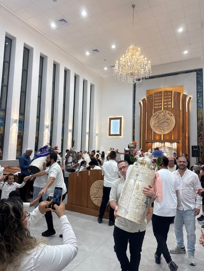 The image depicts a celebratory gathering inside a large, modern synagogue or Jewish community center, located in Beer Yaaqov, Israel.

The scene captures a dynamic moment, likely during a Hachnasat Sefer Torah or Simchat Torah celebration, characterized by individuals carrying multiple Torah scrolls. In the foreground, a woman with brown curly hair, wearing a white long-sleeved shirt, holds a smartphone horizontally, recording the event.

In the mid-ground, numerous men and some children are active participants. A man with a beard, wearing a white shirt and dark pants, is seen carrying a Torah scroll encased in a blue and white decorative mantle, adorned with red flowers, appearing to move rhythmically. To his right, a young boy smiles. Further right, a man wearing glasses, in a light-colored short-sleeved shirt and dark pants, carries a large, intricately decorated silver-cased Torah scroll, which features a pink sash and blue floral accents. He looks towards the camera with a slight smile. Beside him, another man in a white polo shirt and light-wash jeans also smiles directly at the camera. Other men and children, mostly in white shirts and wearing yarmulkes, are visible throughout the space, some carrying additional Torah scrolls or observing. A wooden *bimah* or reading desk is positioned in the mid-ground.

The background features a grand interior architecture. At the far end, a large, ornate wooden Aron Kodesh (Holy Ark) with Hebrew inscriptions and a central circular carving (possibly depicting a lion or other symbol) serves as a focal point. Tall, narrow vertical windows with dark frames line the left wall, extending towards the high ceiling. The walls are white, and the floor is composed of light-colored tiles. A large, multi-tiered crystal chandelier hangs prominently from the white ceiling, which also features recessed lighting and air vents. A framed document with Hebrew text is mounted on the wall to the right of the windows. Stained-glass elements are visible flanking the Aron Kodesh and possibly below the tall windows. The overall lighting is bright, emanating from artificial sources.