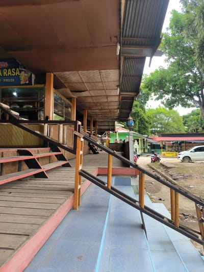 This image captures a long, covered outdoor walkway or veranda of a commercial establishment, likely a restaurant or cafe, set in Bateiiliek, Indonesia. The structure features a wooden plank floor, an elevated platform with steps, and rustic wooden handrails. The building's facade is primarily brown, with large glass windows showcasing what appears to be a food display inside. Above, a corrugated metal roof is visible, with a woven mat-like material lining the underside of the overhang.
Beyond the wooden structure, the ground transitions to blue and light blue tiles, then to an unpaved dirt area. Several vehicles are present: a white car is parked on the dirt, alongside two motorcycles (scooters), one red and one red/black. In the background, there's a light green building with a distinct pointed roof, possibly a mosque or other local structure, and further buildings with red roofs. Lush green trees are abundant, suggesting a tropical environment.
The time of day appears to be daytime, with bright, diffused lighting, indicating either an overcast sky or bright daylight without direct sun. No specific activity or event is taking place; the scene is quiet.
Visible text on a blue banner on the building reads "G DI," "RASA," and "A - BIREUEN," accompanied by a graphic of a drink. Bireuen is a regency in Aceh, Indonesia. Another yellow sign near the white car clearly displays "BATEEILIEK," confirming the specific location context provided. The overall scene conveys a casual, local atmosphere.