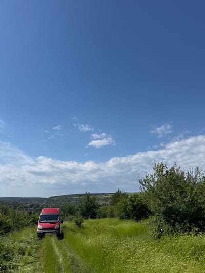 A slightly rotated outdoor scene features a red van positioned on a grassy track, surrounded by dense green vegetation and trees. The sky occupies a significant portion of the image, transitioning from a clear blue to wispy clouds near the horizon. The environment suggests a rural or agricultural setting, with hints of habitation visible in the distance. Although the image is rotated, the details of the van, the surrounding vegetation, and the sky are clearly visible. There is no evidence indicating that this scene is within the city of Topchii, Bulgaria, or any other specific location. There are no visible people or actions taking place.