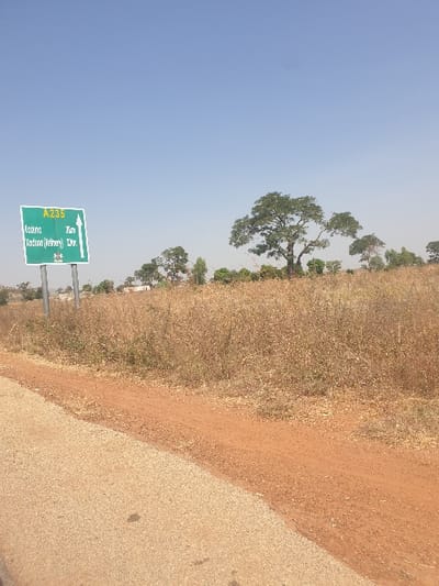 A static, outdoor scene captured under a clear blue sky, located on a road within or immediately adjacent to Kujama, Nigeria. The foreground presents a light-colored paved road surface on the lower left, adjacent to a reddish-brown dirt shoulder. Beyond the shoulder, a wide expanse of dry, golden-brown vegetation dominates the middle ground. On the left, a green rectangular road sign, mounted on two light-colored metallic poles, displays white text: "A.235" at the top, followed by "Kaduna 3km" and "Kaduna (Refinery) 12km," both accompanied by upward-pointing arrows. The background features several trees with green foliage, including a large, prominent tree situated towards the right of the frame. No individuals, animals, or vehicles are present. The environment suggests a dry seasonal period.