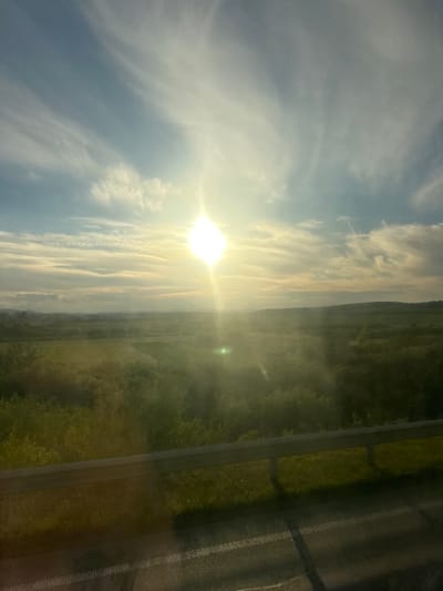 A bright, sunlit landscape is captured in this image, likely taken from a moving vehicle. The sun is intensely bright, positioned high in the sky and radiating light that creates a glare and lens flare effect across the scene. Wispy clouds streak across a pale blue sky. Below the sky is a vast, green expanse, appearing to be rural or agricultural land, with rolling hills in the distance. A dark line, possibly a guardrail, runs horizontally across the lower portion of the image, suggesting a roadside perspective. The foreground shows asphalt with white markings, confirming the presence of a road. The overall impression is one of a sunny day in a natural, open environment. The location is identified as Pidbirtsi, Ukraine.