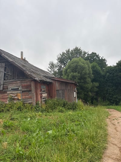 The image depicts an old, weathered wooden house in a rural setting, likely in Ashmyany, Belarus. The house is in disrepair, with peeling red paint, a decaying roof, and boarded-up windows. Overgrown grass and weeds surround the structure, and a dirt path curves away into the distance. Lush green trees form a dense backdrop to the scene. The sky is overcast, contributing to a somber and neglected atmosphere. There are no people or animals visible in the image.