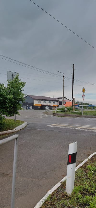 The image depicts a view of a street in Agapovka, Russia, under an overcast sky. In the foreground, a wet asphalt road with a white and red striped marker pole is visible. A crosswalk is painted on the road. To the left, a metal fence runs along the side of the road, bordered by green grass.
In the middle ground, a modern-looking building with a dark grey facade, white window frames, and a brown roof stands. Orange signage is present on the lower section of the building. To the right of this building, other structures, including a gas station with visible pumps and blue awnings, are present. Power lines and utility poles are visible throughout the scene, including a prominent wooden pole with insulators. The overall environment appears to be a commercial or mixed-use area. There are no people visible in the image.